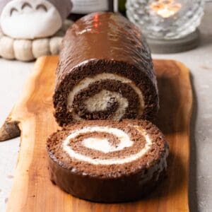 Chocolate Swiss roll cake with cream cheese filling is covered with dark chocolate glaze and served on a wooden serving board. One piece has been cut off and is lying on the same board.