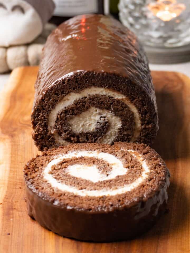 Chocolate Swiss roll cake with cream cheese filling is covered with dark chocolate glaze and served on a wooden serving board. One piece has been cut off and is lying on the same board.