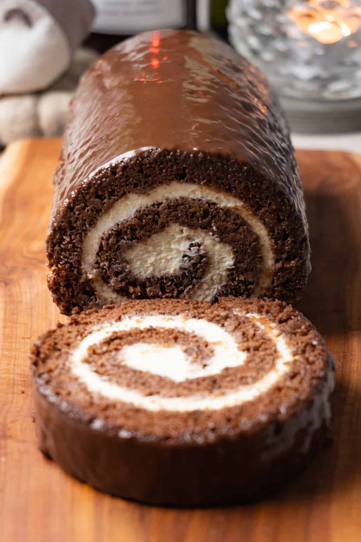 Chocolate Swiss roll cake with cream cheese filling is covered with dark chocolate glaze and served on a wooden serving board. One piece has been cut off and is lying on the same board.
