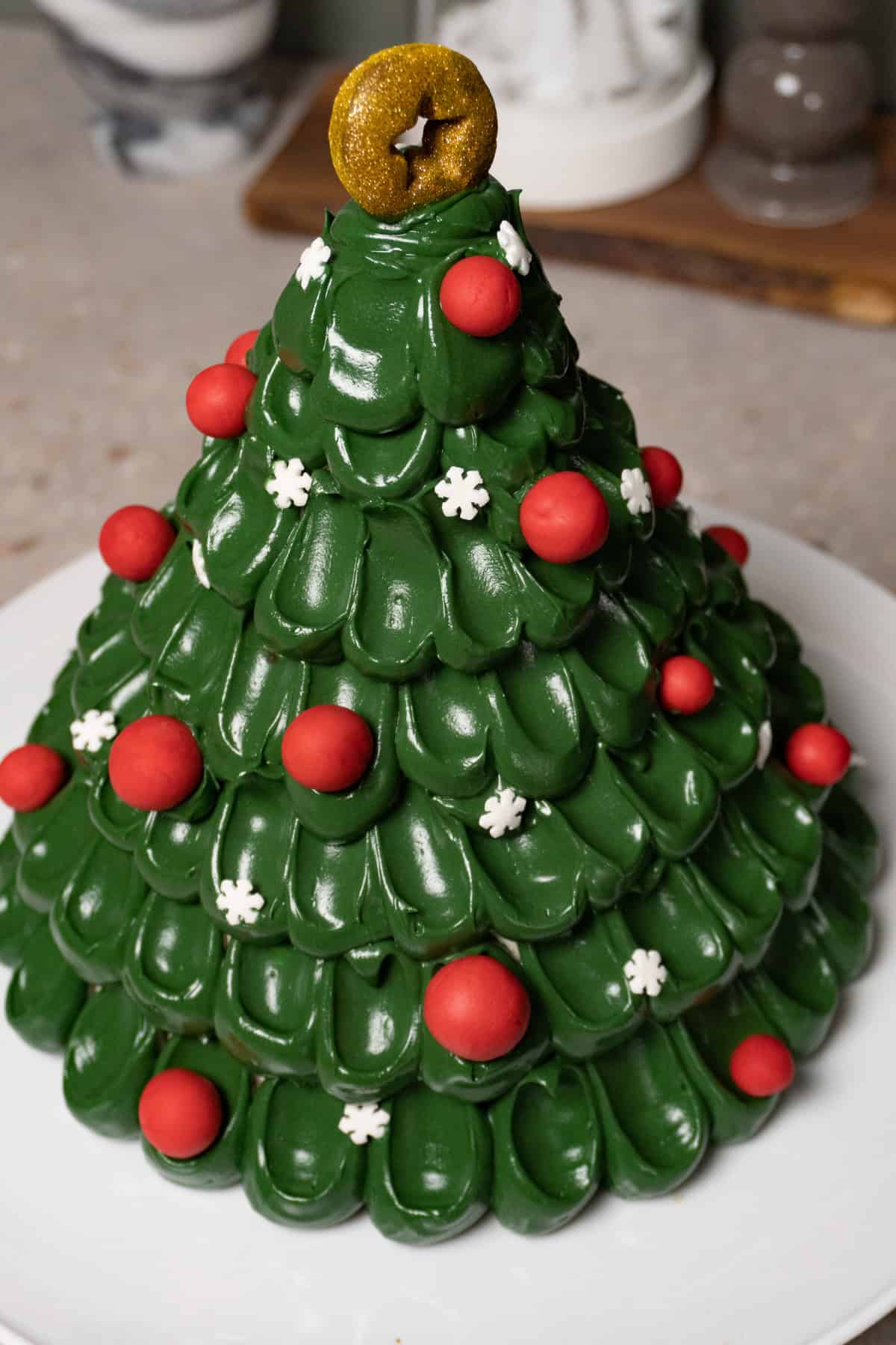 A green Christmas tree cake decorated with red baubles, snowflake sprinkles, and a gold star topper sits on a white cake stand.