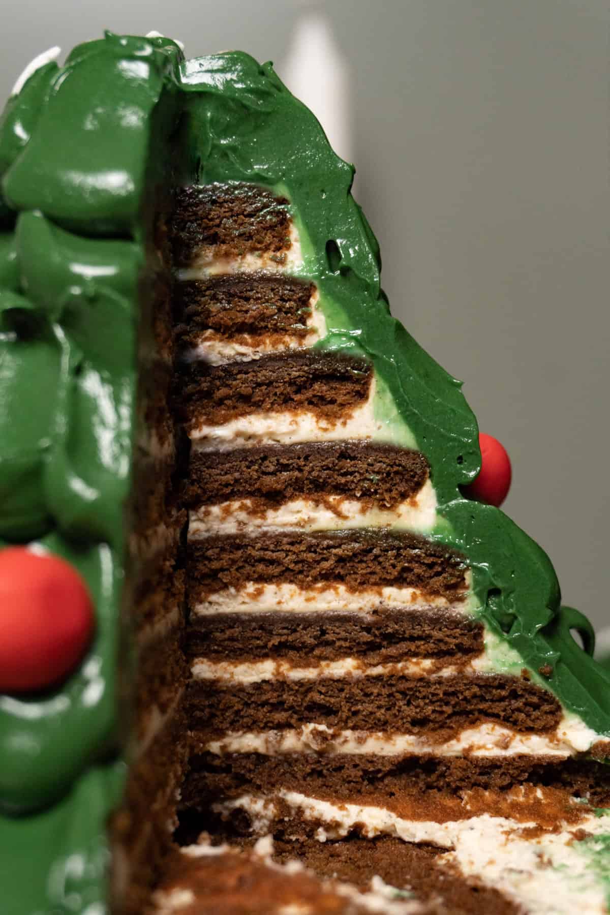 A green Christmas tree cake with red baubles and snowflake sprinkles; one piece has been taken, revealing thin chocolate cake layers and white cream filling.