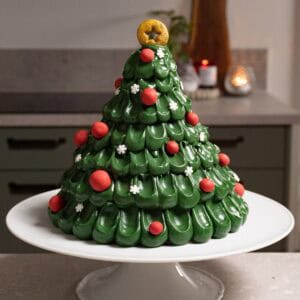 A green Christmas tree cake decorated with red baubles, snowflake sprinkles, and a gold star topper sits on a white cake stand.