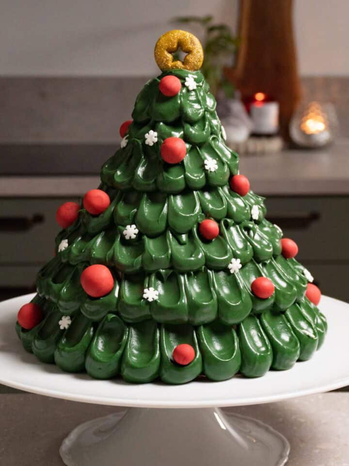 A green Christmas tree cake decorated with red baubles, snowflake sprinkles, and a gold star topper sits on a white cake stand.