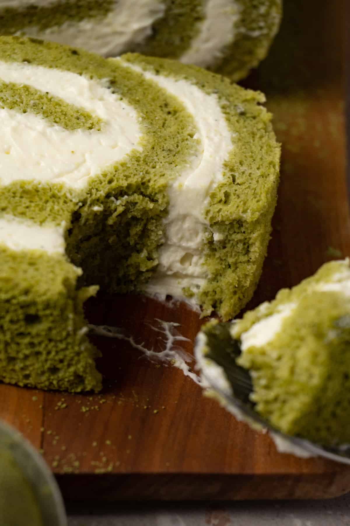 A piece of matcha Swiss roll cake with cream cheese filling on a wooden board, with one bite taken using a spoon.