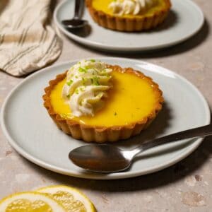 A mini lemon tart with white cream topping sits on a plate with a silver spoon.