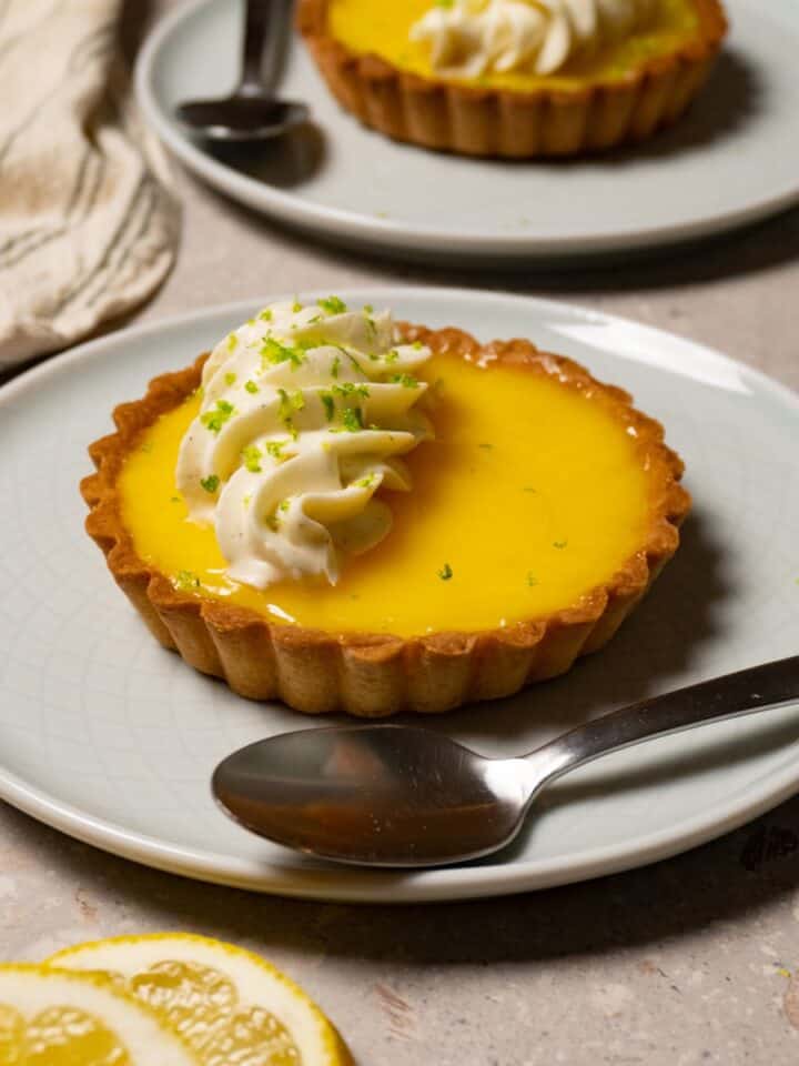 A mini lemon tart with white cream topping sits on a plate with a silver spoon.