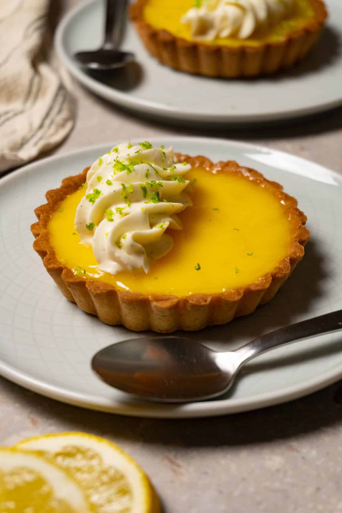 A mini lemon tart with white cream topping sits on a plate with a silver spoon.