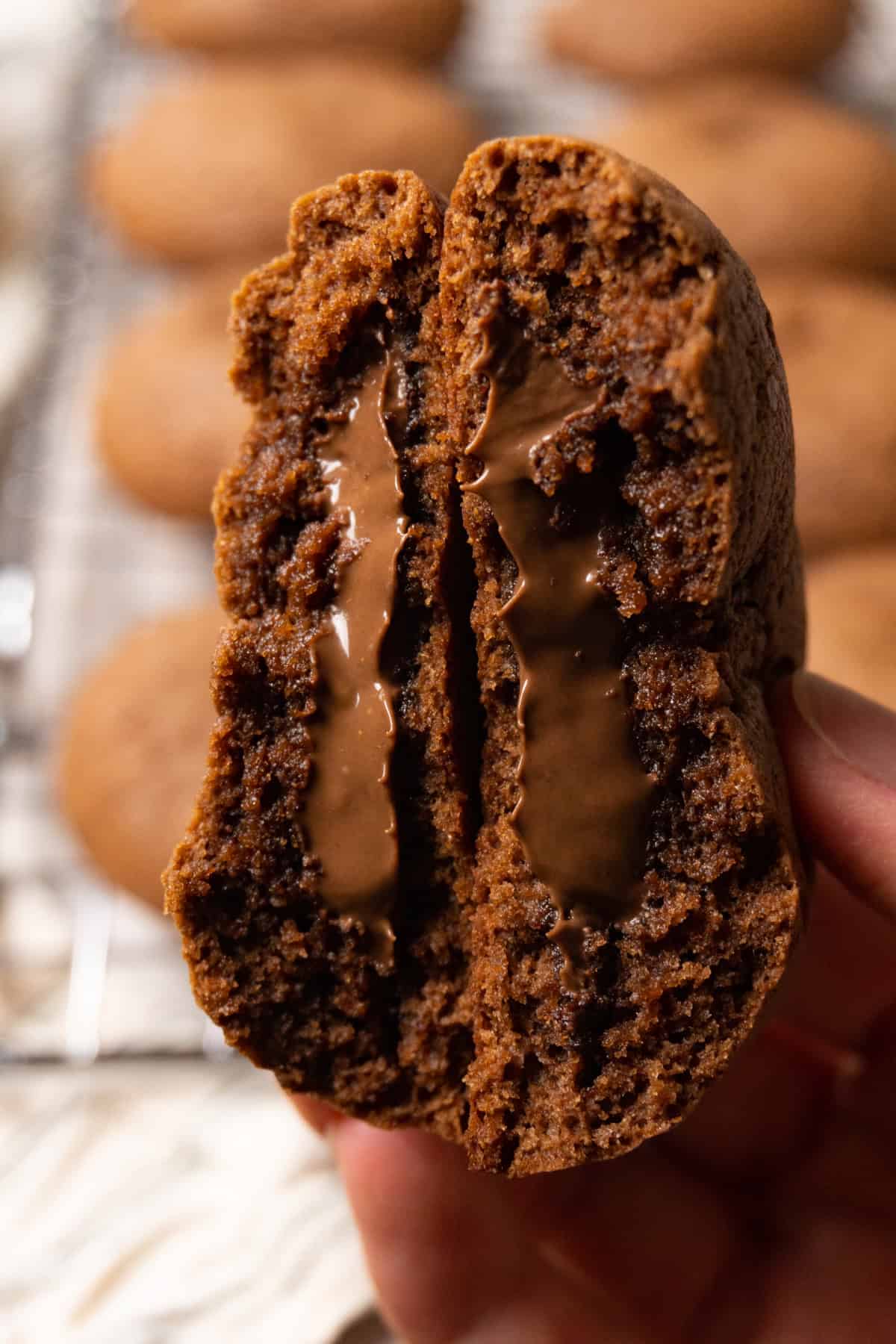 A hand is holding a chocolate cookie broken in half, with chocolate filling oozing from the center.