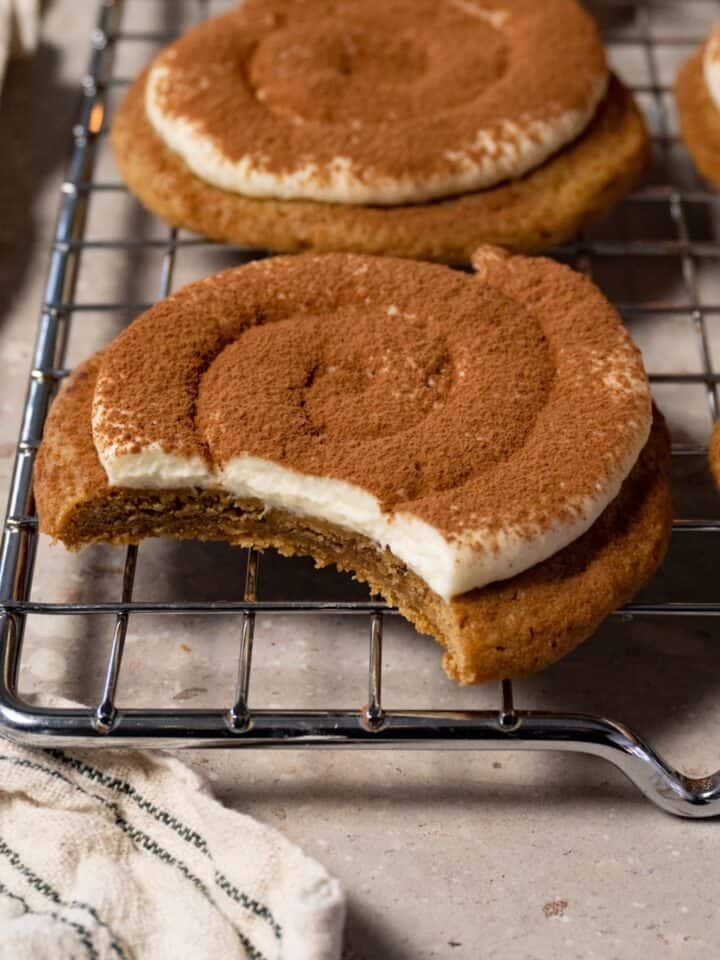 A cookie topped with cream and dusted with cocoa powder rests on a metal tray, with one bite taken.