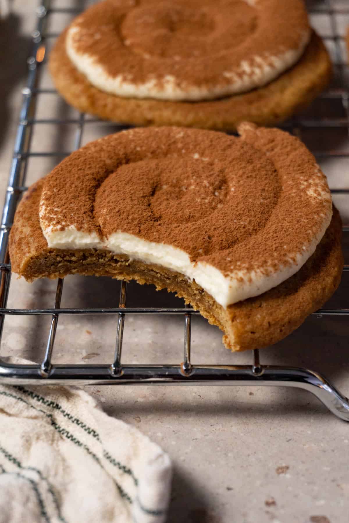 A cookie topped with cream and dusted with cocoa powder rests on a metal tray, with one bite taken.