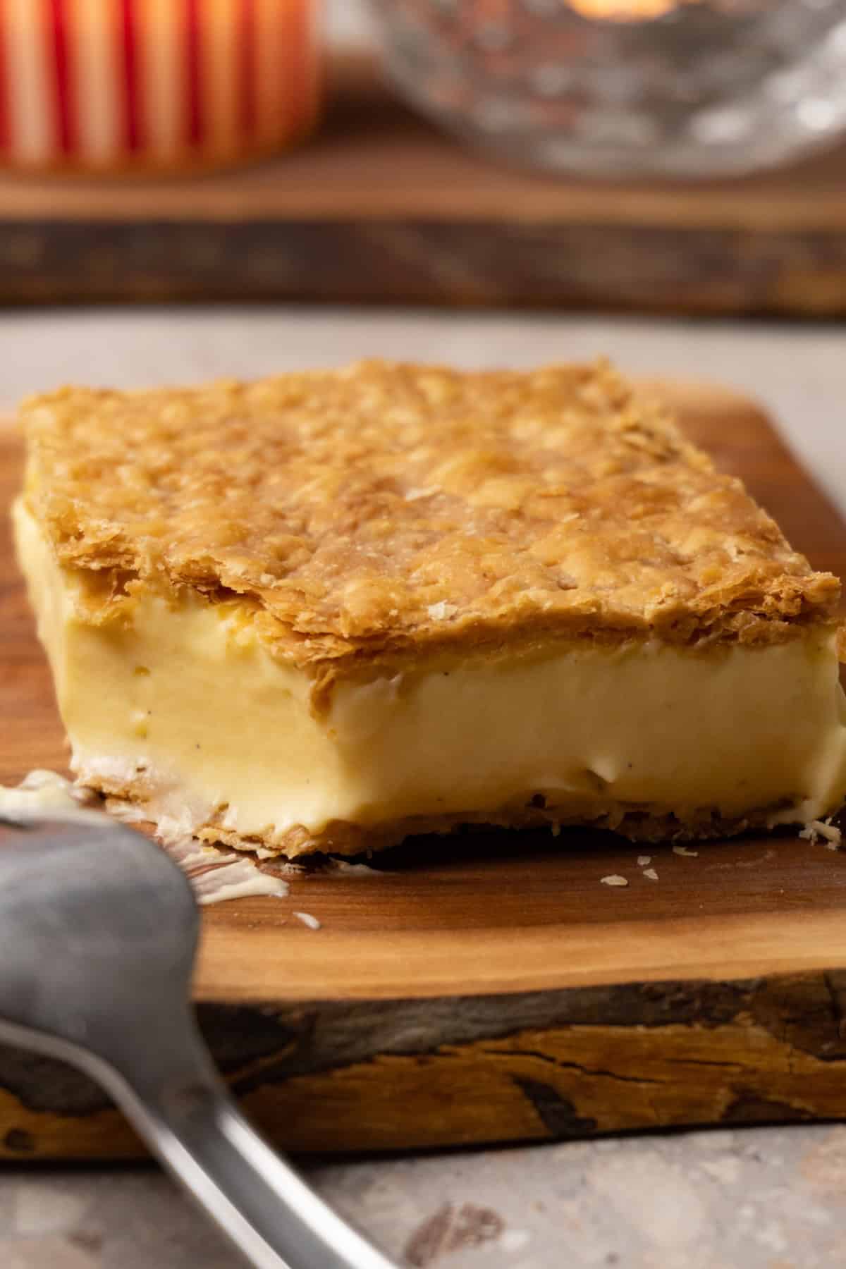 slice of pastry made with two layers of puff pastry and vanilla cream in the middle lies on a wooden board, with one bite taken out of it.