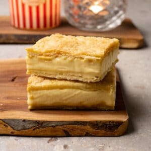 Two slices of pastry made with layers of puff pastry and vanilla cream in the middle are stacked on a wooden board.