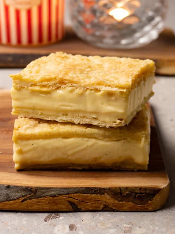 Two slices of pastry made with layers of puff pastry and vanilla cream in the middle are stacked on a wooden board.
