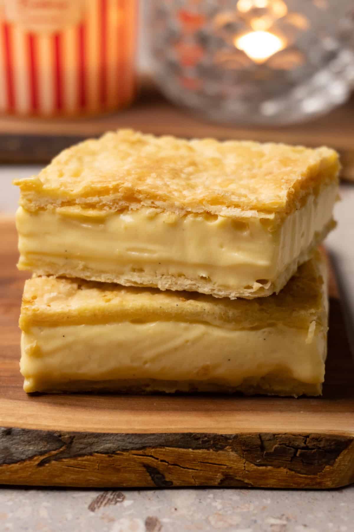 Two slices of pastry made with layers of puff pastry and vanilla cream in the middle are stacked on a wooden board.