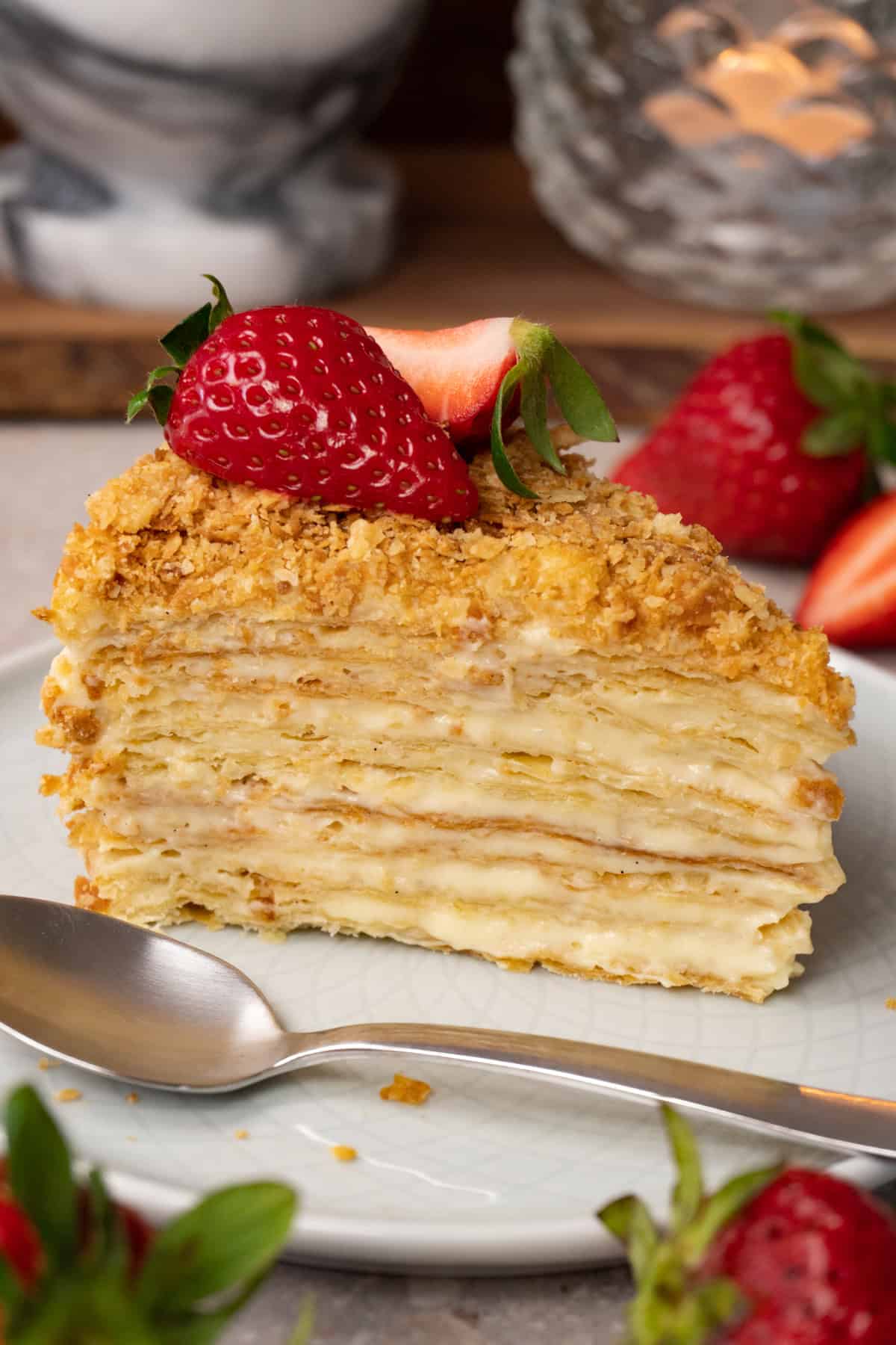 A piece of cake made with puff pastry layers and pastry cream in between, topped with fresh strawberries, is served on a plate with a silver spoon.