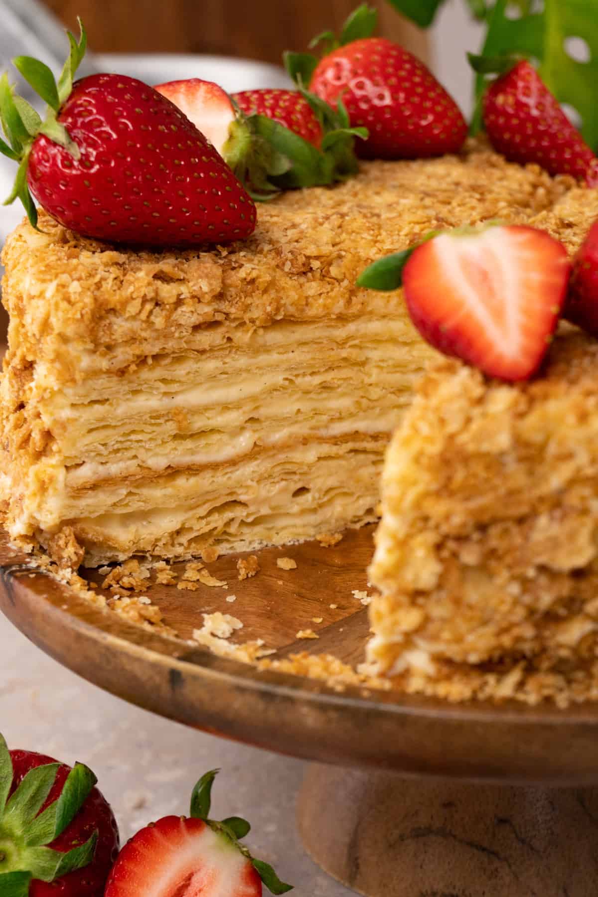 A cake made with puff pastry layers and pastry cream in between, topped with fresh strawberries, is served on a wooden cake stand, with one piece already taken.