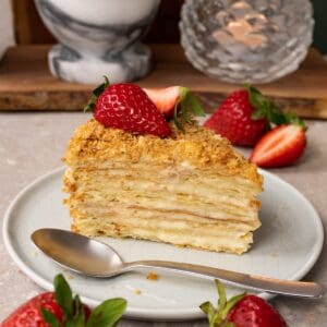 A piece of cake made with puff pastry layers and pastry cream in between, topped with fresh strawberries, is served on a plate with a silver spoon.