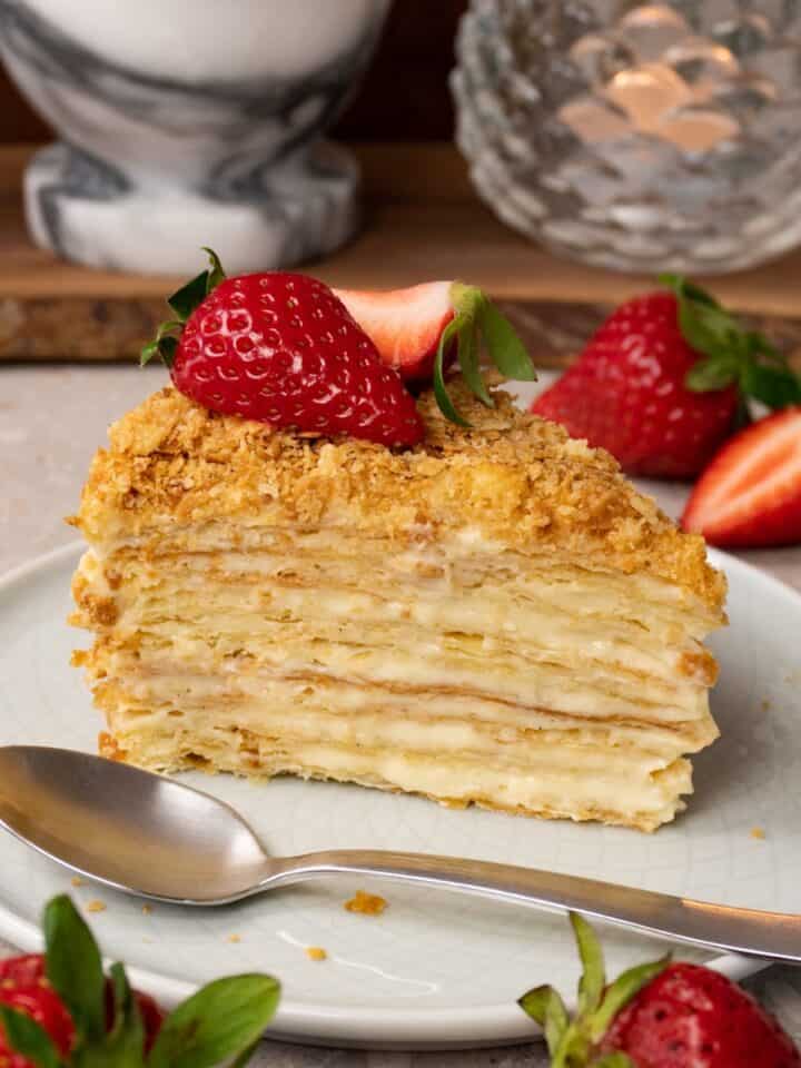 A piece of cake made with puff pastry layers and pastry cream in between, topped with fresh strawberries, is served on a plate with a silver spoon.