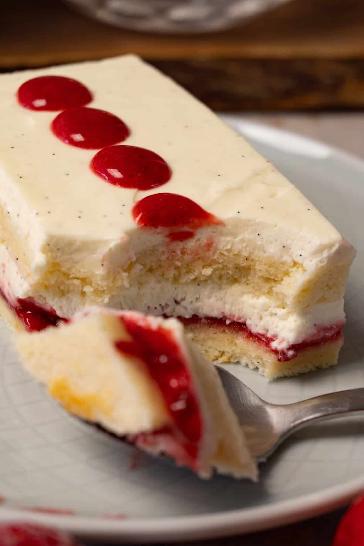 A rectangular piece of cake with white cream frosting and strawberry jam filling is on a round plate with a silver spoon. One bite has been taken.
