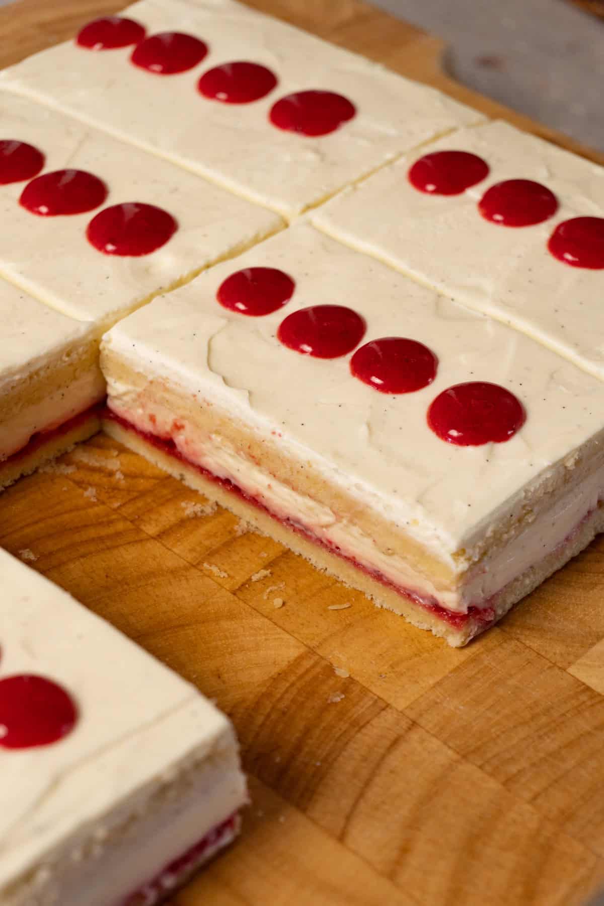 A rectangular cake with white cream and strawberry jam filling on a wooden board. One rectangular piece has been cut out.