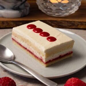 A rectangular piece of cake with white cream frosting and strawberry jam filling is on a round plate with a silver spoon.