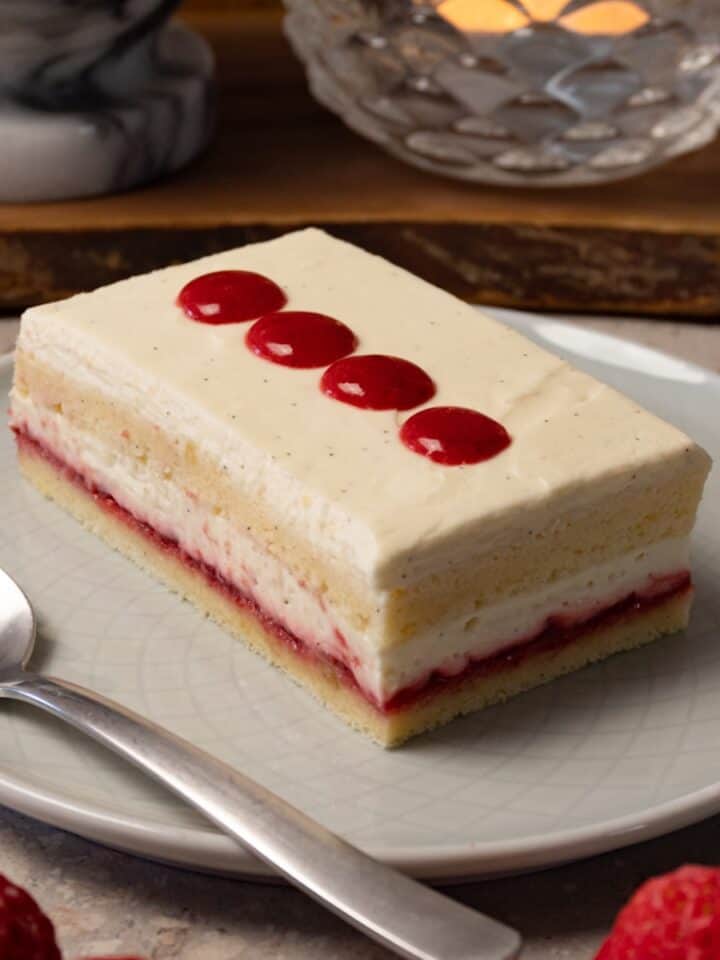 A rectangular piece of cake with white cream frosting and strawberry jam filling is on a round plate with a silver spoon.