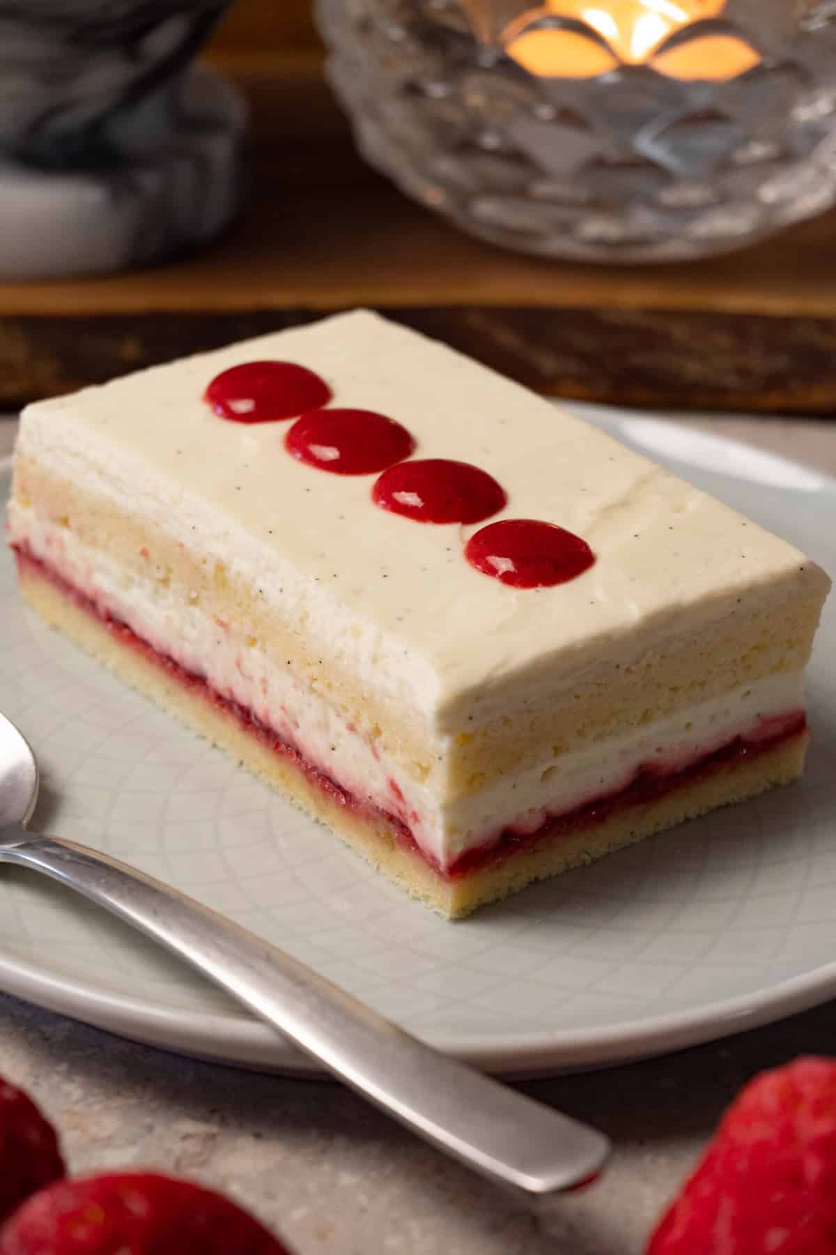 A rectangular piece of cake with white cream frosting and strawberry jam filling is on a round plate with a silver spoon.